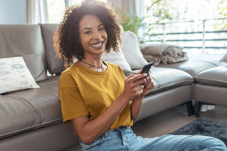 Happy mature woman using smartphone while relaxing sitting on the floor looking at camera at home.